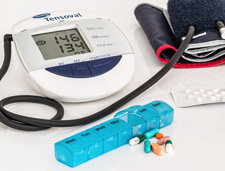 Image of blood pressure reader, weekly pill case, and pack of medicine sitting on white table.