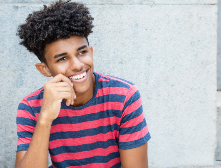 image of a kid smiling, hand on cheek, sitting against a concrete wall